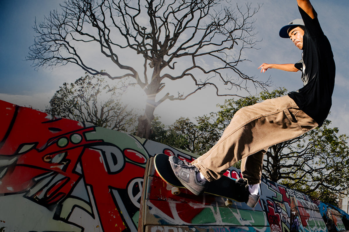 Skateboarder performing a trick on a graffiti-covered ramp outdoors, wearing a black T-shirt, brown pants, and gray skate shoes.