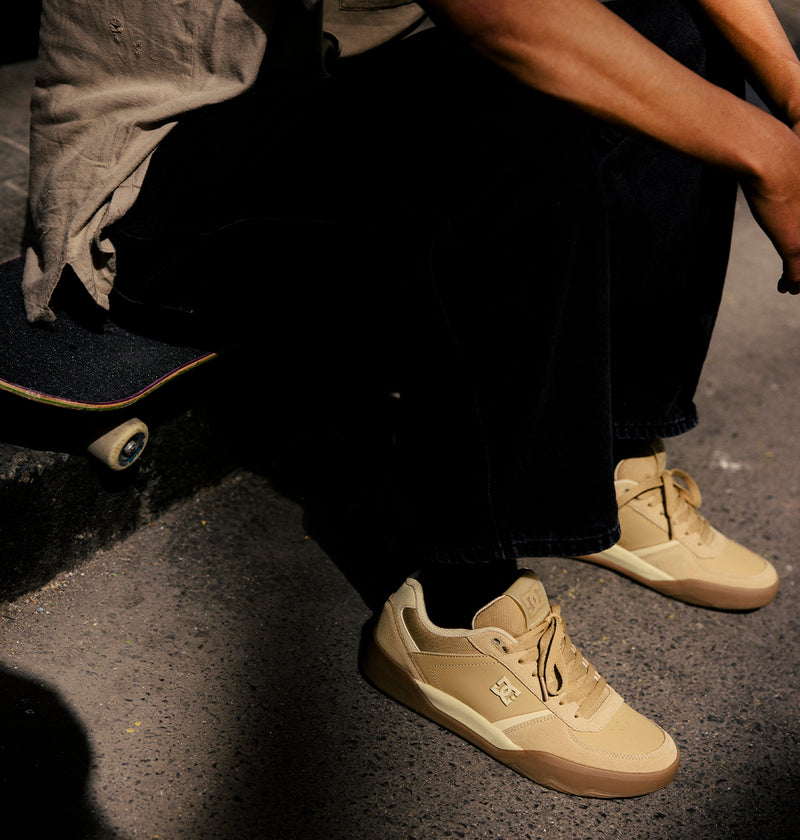 A person sits on a skateboard, wearing black pants and the DC Shoes Men's Fractal S Shoes in beige, as sunlight casts shadows on the ground—showcasing street-ready style and performance-driven comfort.