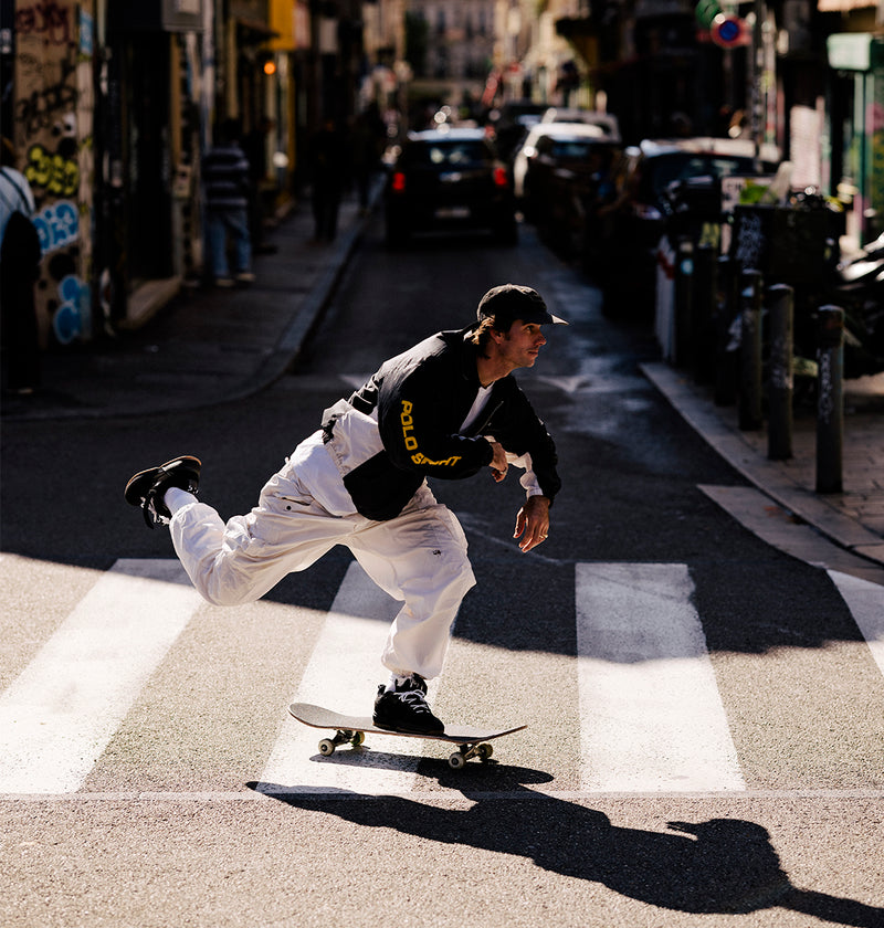 Skateboarder performing a trick on a city street with cars and buildings in the background.