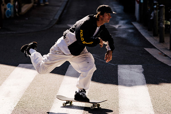 A person wearing a black cap, black sweatshirt, and white pants is skateboarding across a sunlit street, one foot on the skateboard and the other pushing off the ground.
