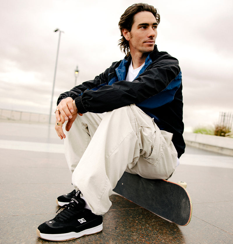 Channeling DC Shoes heritage, a man in a dark jacket and light cargo pants sits on a skateboard at an urban skate park, wearing Men's JS-2 Shanahan Pro Skate Shoes—resting his arms on his knees and gazing sideways beneath a cloudy sky.