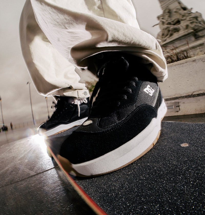 A person sports white pants and black DC Shoes Men's JS-2 Shanahan Pro Skate Shoes, inspired by John Shanahan, while standing on a skateboard outdoors. Sunlight gleams off the ground with a statue and streetlights behind.