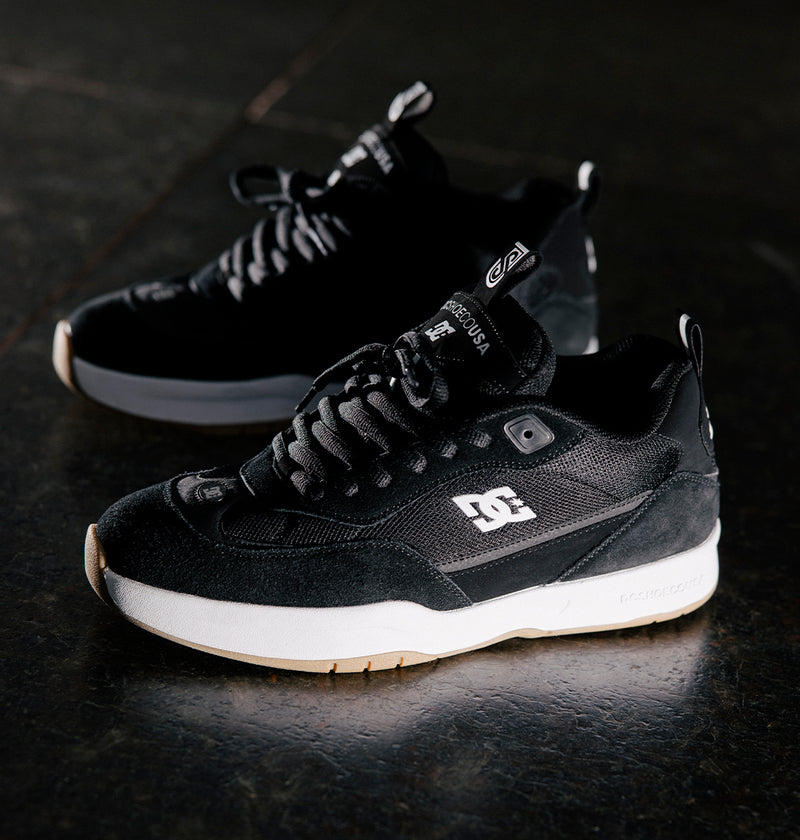 A pair of Men's JS-2 Shanahan Pro Skate Shoes by DC Shoes in black with white soles and branding, inspired by DC heritage, are displayed on a dark surface with one shoe slightly ahead of the other.