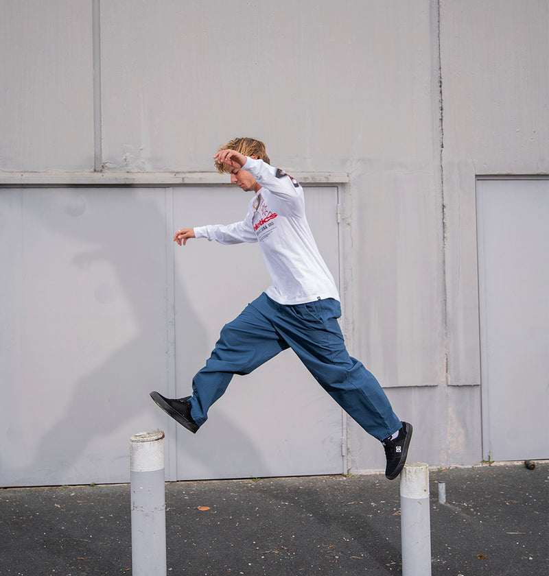Wearing the DC Shoes Men's Slathletic Heritage Long Sleeve Shirt, a man jumps between two white posts in front of a gray concrete wall.