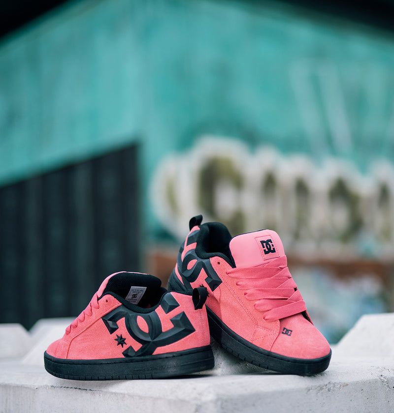 Two pairs of Men’s Court Graffik Beveled Special Edition Shoes by DC Shoes in pink and black are displayed outdoors on a white surface, highlighting their puffy silhouette and bold DC branding, with a blurred teal and graffiti-covered wall in the background.
