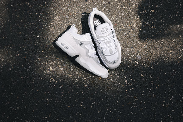 A pair of white DC brand sneakers are placed on a sunlit patch of asphalt, with some shadow and light patterns on the ground around them.