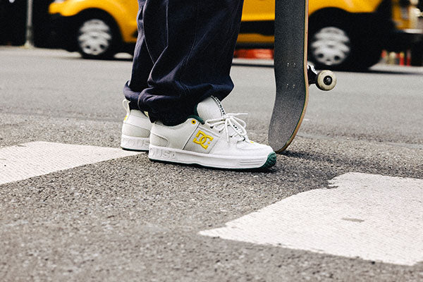 White sneakers with a DC logo on a street with a skateboard and blurred vehicle in the background