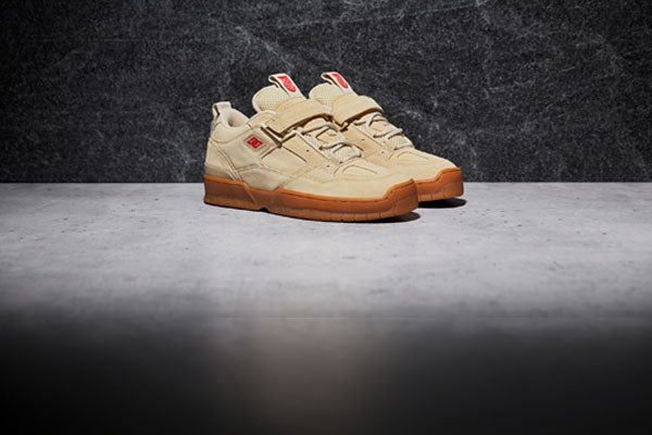 A pair of beige sneakers with gum soles are positioned side by side on a gray, concrete-like surface with a dark textured background.