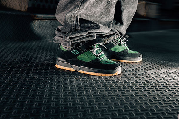 Green and black sneakers worn with gray pants on a textured floor.