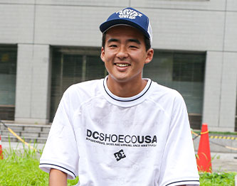 Shintaro Kongo wearing a white t-shirt with 'DC SHOE CO. USA' logo and a blue cap, sitting outdoors.