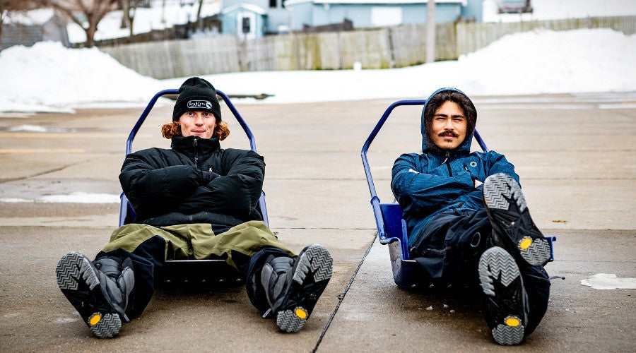 Two people sitting on blue and black sleds on a snowy street.